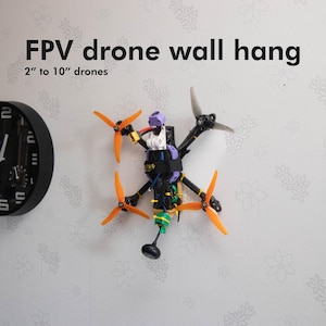 Puede incluir: Un pequeño dron con hélices naranjas y un reloj blanco y negro en una pared blanca. El texto "FPV drone wall hang 2" to 10" drones" está impreso en la pared.