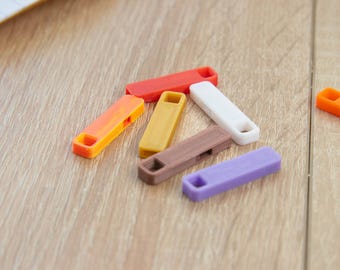 Op de afbeelding: Zes kleurrijke plastic sleutelhangers in verschillende tinten oranje, geel, bruin, wit en paars. De sleutelhangers zijn in een halve cirkel op een houten oppervlak gerangschikt.