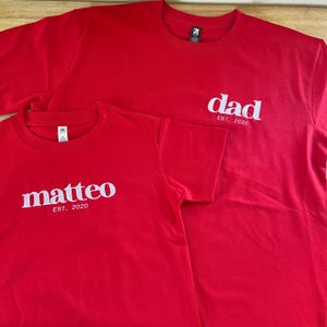 May include: Two red short-sleeved t-shirts with white text. The larger shirt features the word "dad" above "EST. 2020". The smaller shirt features the name "matteo" above "EST. 2020". Both shirts have a crew neckline and a visible interior brand label.