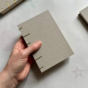 Op de afbeelding: Een handgemaakt grijs papieren notitieboek met zichtbare groene stiksels langs de rug. Het notitieboek wordt in een hand gehouden, wat het gedetailleerde vakmanschap laat zien. Extra notitieboeken zijn zichtbaar op de achtergrond.