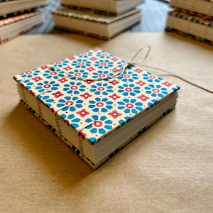 Może przedstawiać: Ręcznie robiony notatnik z niebieskim i czerwonym kwiatowym wzorem na kremowym tle. Notatnik jest kwadratowy, z zapięciem na klapę i sznurkiem. Inne notatniki są ułożone w stosy w tle.