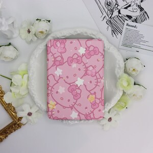 May include: A pink notebook featuring a repeating pattern of Hello Kitty faces, bows, and stars. The notebook is surrounded by white flowers and a decorative gold frame. The background is white.