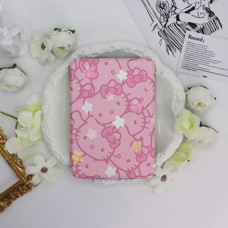 May include: A pink notebook featuring a repeating pattern of Hello Kitty faces, bows, and stars. The notebook is surrounded by white flowers and a decorative gold frame. The background is white.
