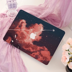 May include: Laptop cover featuring a vibrant design of red clouds against a dark blue starry sky. A white Apple logo is prominent, with a white arrow and birds. The cover is on a white surface with pink fabric and flowers.