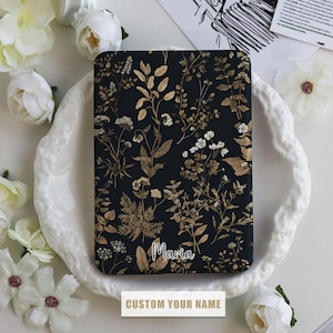 May include: A black and gold floral pattern phone case with the name "Maria" printed on it. The case is on a white plate with flowers.