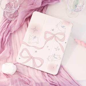 May include: A light pink tablet case featuring a repeating pattern of flying bunny illustrations, pink bows, and stars. The case is open, revealing the screen. The background includes a pink scarf, a glass, and a candle.