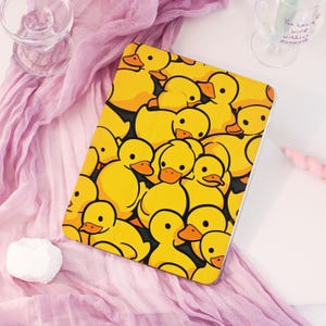 May include: A yellow tablet case featuring a repeating pattern of cartoon ducks with orange beaks and black eyes. The ducks are set against a black background. The case is on a pink fabric.