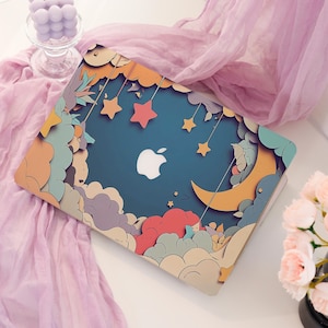 May include: A laptop with a whimsical design featuring a blue background with a white Apple logo. The cover is decorated with colorful clouds, stars, and a crescent moon. The design has a dreamy, cartoonish aesthetic.