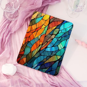 May include: A rectangular tablet case with a stained glass design. The case features a vibrant pattern of abstract shapes in shades of orange, blue, green, and brown, creating a colorful mosaic effect. The case is on a pink surface.
