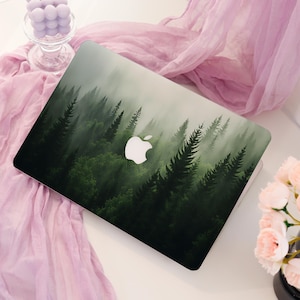 Op de afbeelding: Een laptop met een skin in bosthema met donkergroene dennenbomen en een mistige, nevelige sfeer. Het iconische witte Apple-logo is in het midden van het deksel geplaatst en steekt af tegen het diepgroene gebladerte en de bosachtergrond.