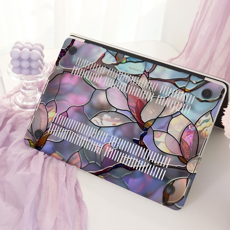 May include: A laptop case with a stained-glass floral design in shades of purple, pink, and blue. The case features a pattern of stylized flowers and leaves, with white vent lines. The case is on a white surface.