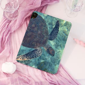 May include: A rectangular tablet case featuring a detailed image of a sea turtle swimming in turquoise water. The turtle's shell is a mosaic of dark brown and green, with patterned flippers. The case is on a pink fabric.