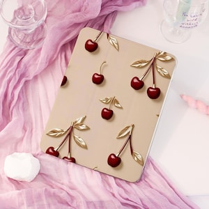 May include: A beige tablet case featuring a repeating pattern of 3D cherries with gold-colored leaves. The cherries are a deep red color. The case is on a pink fabric surface.