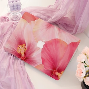 Peut inclure: Un ordinateur portable avec un motif floral. La coque présente de grands pétales d'hibiscus roses et rouges avec des étamines jaunes. Le logo Apple est visible sur la coque. Le fond comprend une écharpe lavande et un petit bouquet de fleurs.