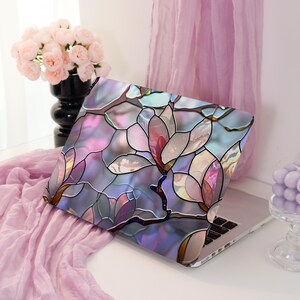 May include: A laptop with a decorative cover featuring a stained-glass floral design in shades of pink, purple, and blue. The laptop is open, displaying the keyboard. A vase of pink flowers and a lavender candle are in the background.
