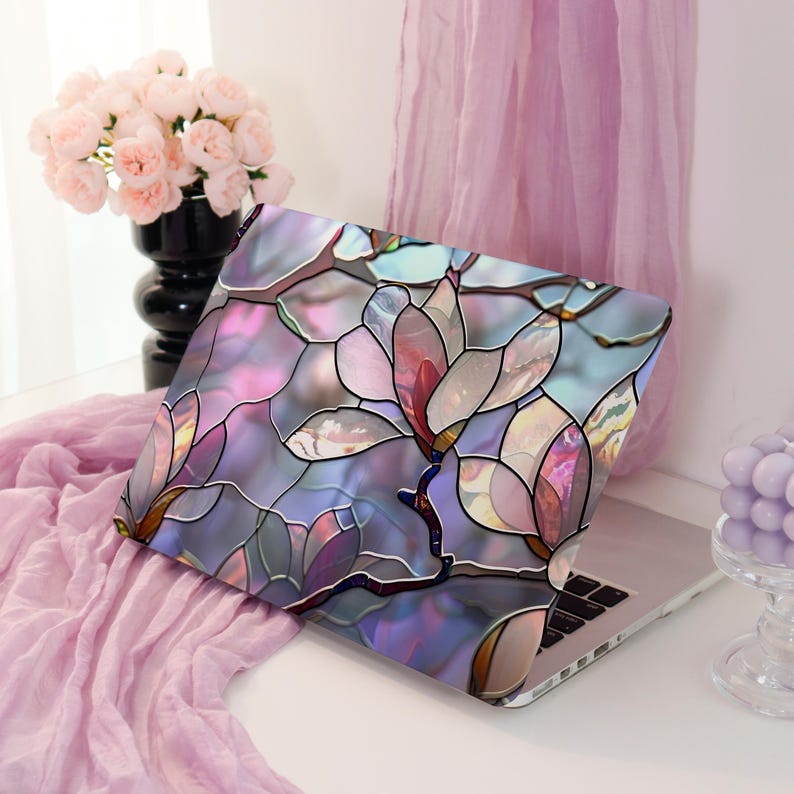 May include: A laptop with a decorative cover featuring a stained-glass floral design in shades of pink, purple, and blue. The laptop is open, displaying the keyboard. A vase of pink flowers and a lavender candle are in the background.