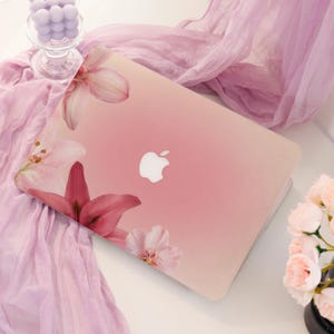 May include: A laptop with a soft pink gradient finish featuring a floral design of lilies and cherry blossoms on the cover. The center of the lid displays a white Apple logo. The laptop is positioned on a surface with light purple fabric.
