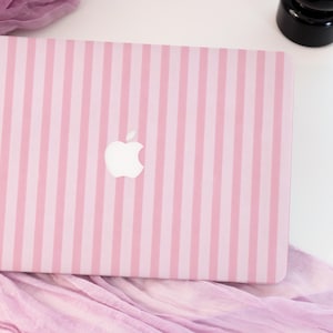 以下が含まれることがあります： ピンクと白のストライプ柄のノートパソコンカバー。Appleのロゴ入り。カバーは明るいピンク色で、少し濃いピンク色の縦縞模様。Appleのロゴは白で、カバーの中央に配置されています。
