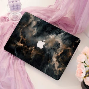 May include: A laptop featuring a black and gold abstract design with swirling patterns and a white Apple logo. The laptop is placed on a white surface, with a pink cloth and flowers in the background.