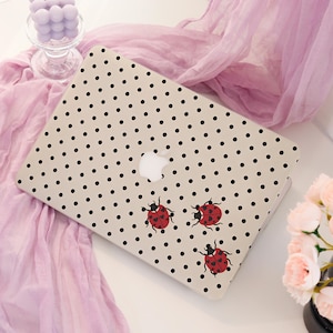 May include: A laptop skin featuring a beige background with a repeating pattern of small black polka dots and three red ladybugs with black spots. The center of the laptop lid displays a white Apple logo cutout, showing the device brand underneath.