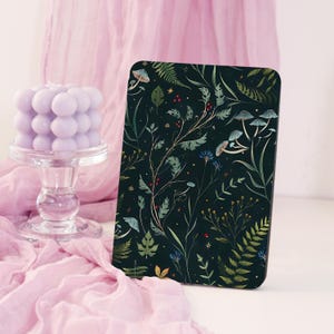 May include: A dark green rectangular object with a botanical design featuring mushrooms, leaves, and flowers. A light purple bubble candle sits on a clear glass pedestal, with pink fabric in the background.