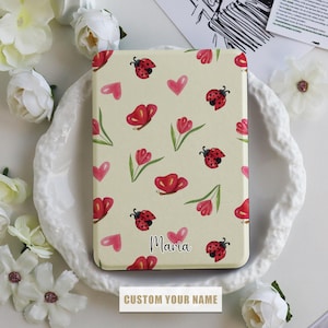 Pode incluir: Uma capa de cor creme para Kindle Paperwhite com um padrão floral que inclui joaninhas vermelhas, corações cor-de-rosa e folhas verdes. O texto "Maria" está impresso na capa.