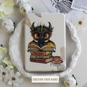 Può includere: Una custodia per e-reader bianca e marrone con un drago di cartone animato che indossa gli occhiali e tiene un mucchio di libri. I libri hanno il testo "read books be kind stay weird". La custodia ha anche una tazza di caffè e il nome "Maria" scritto su di essa.