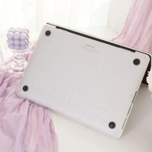 May include: A white MacBook Pro laptop case with black accents. The case has a smooth surface with ventilation slots and four black rubber feet. The laptop is resting on a white surface with a lavender fabric and a decorative glass candle holder in the background.