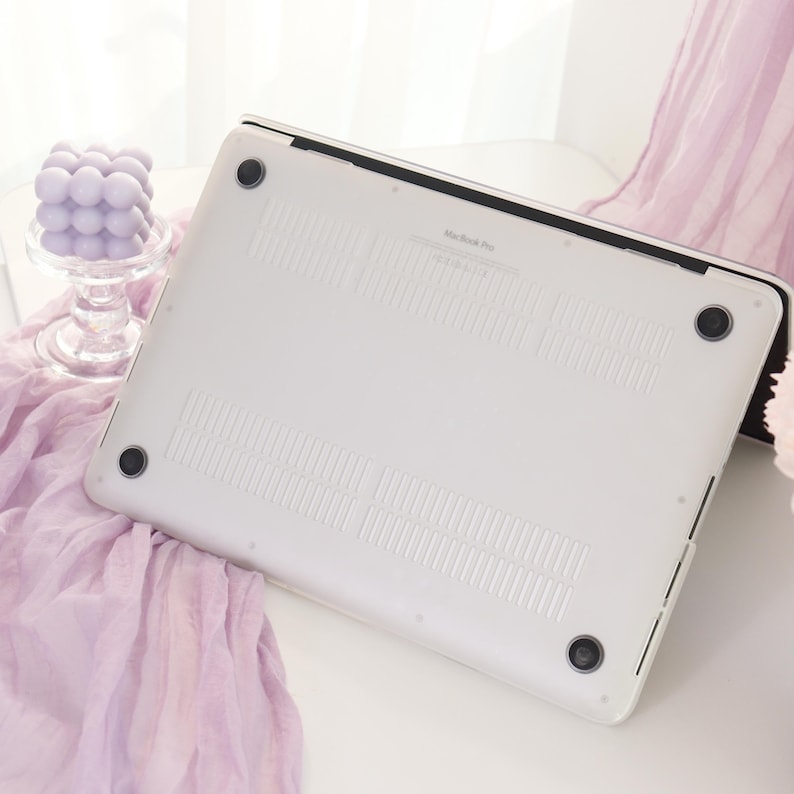 May include: A white MacBook Pro laptop case with black accents. The case has a smooth surface with ventilation slots and four black rubber feet. The laptop is resting on a white surface with a lavender fabric and a decorative glass candle holder in the background.