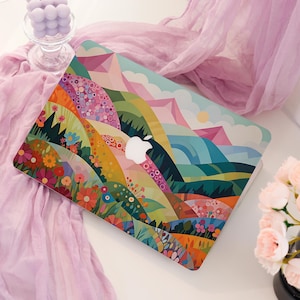 Op de afbeelding: Een laptopskin met een levendige, veelkleurige landschapsillustratie met glooiende heuvels, gestileerde bloemen en bergtoppen. Het ontwerp bevat een palet van roze, oranje, geel, groen en blauw, gecentreerd rond een wit Apple-logo.