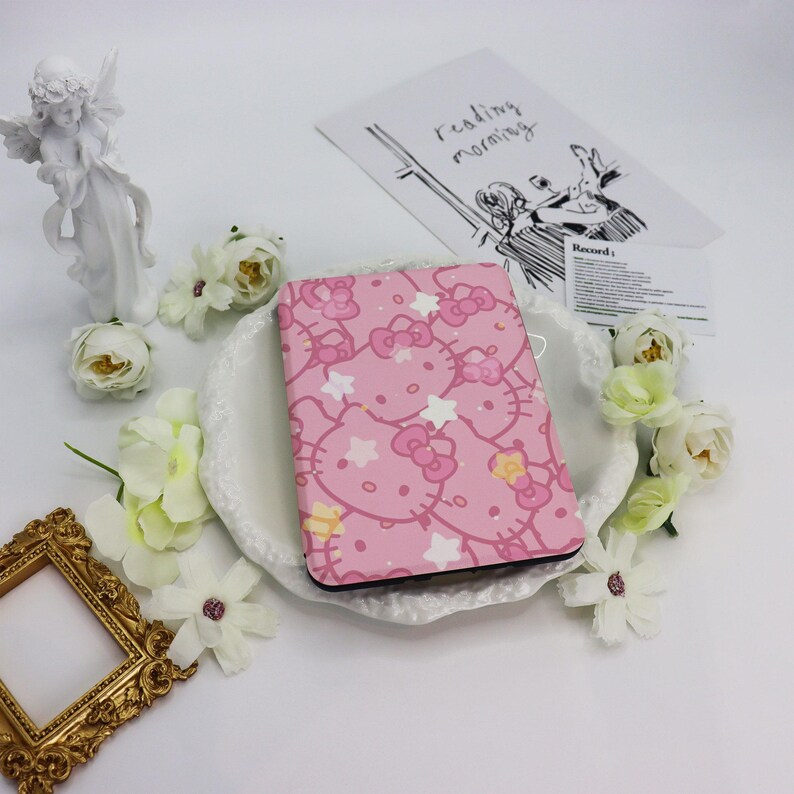 May include: A pink Hello Kitty-themed notebook with a repeating pattern of the character, bows, and stars. The notebook is surrounded by white flowers and a small gold picture frame. A drawing with the words "reading morning" is in the background.