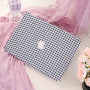 May include: A laptop with a blue and white striped pattern. The laptop has a white Apple logo on the cover. The background includes a pink scarf, a glass candle holder with purple candles, and a bouquet of pink flowers.