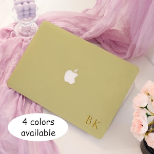 May include: A light green laptop with the Apple logo. The laptop has the initials "BK" in gold. Text on the image says "4 colors available". The laptop is on a white surface with a pink cloth and flowers in the background.
