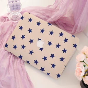 May include: A beige laptop case with a pattern of navy blue stars. The case has a white Apple logo in the center. The laptop case is on a white surface with a pink scarf and flowers in the background.