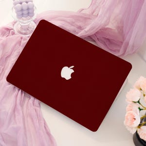 May include: A burgundy laptop with a white Apple logo on the lid. The laptop is closed and positioned on a white surface, with a soft, pink fabric draped nearby. A glass vase with purple decorative balls is visible.