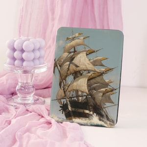 May include: A rectangular decorative panel with a vintage painting of a tall sailing ship. The ship has billowing sails on a choppy sea under a light blue sky. The panel is set against pink fabric and a lavender bubble candle on a glass stand.