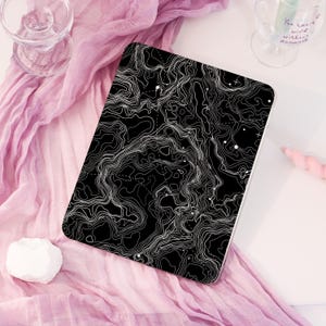 May include: A black and white tablet case featuring a topographic map design. The case has a white border and is set against a pink fabric background. The design includes white contour lines and scattered white dots.