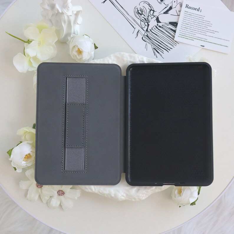 May include: Gray tablet case with a built-in hand strap, open to reveal a black interior. The case is surrounded by white flowers and a decorative card with the word "Record;" printed on it.