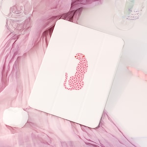 Könnte beinhalten: Weißes Tablet-Case mit einem rosa Geparden-Design mit sternförmigen Punkten. Der Gepard ist in sitzender Position dargestellt. Das Case liegt auf einer rosa Oberfläche mit einem rosa Schal, einem Glas und einem weißen Deko-Objekt.