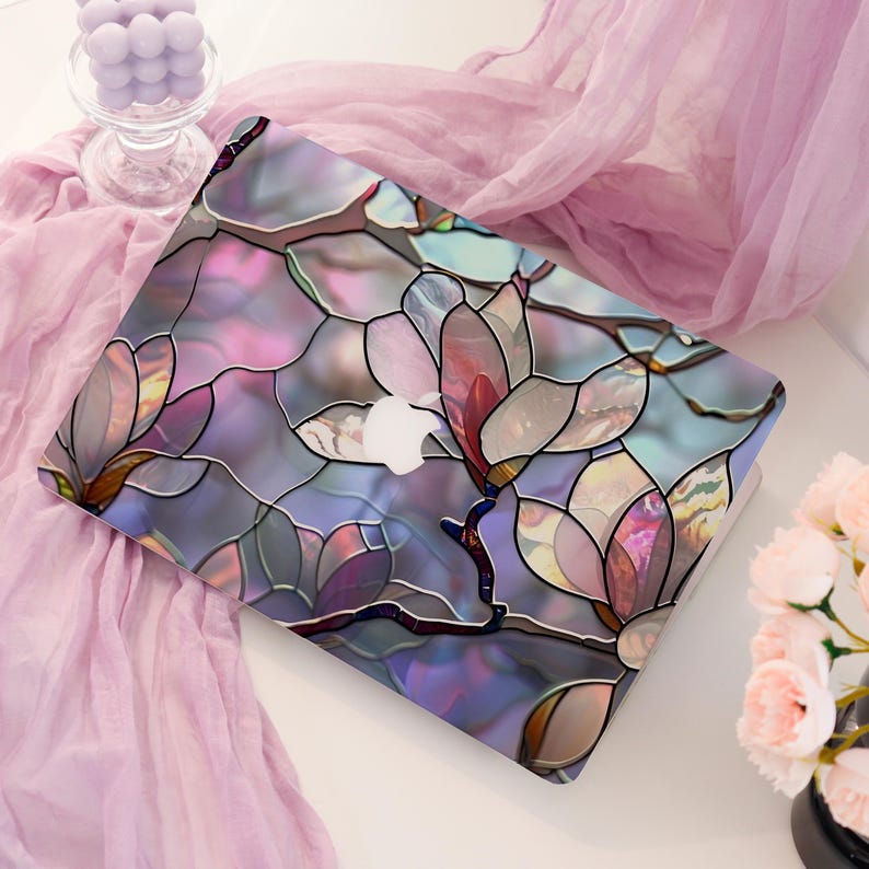 May include: A laptop with a floral design in shades of pink, purple, and blue. The design resembles stained glass, with a white Apple logo visible. The laptop is on a white surface, with a pink scarf and flowers in the background.