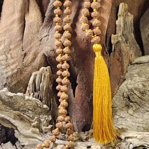 May include: A long necklace featuring numerous small, round, brown, textured rudraksha beads strung on a bright yellow cord. The necklace ends with a long, vibrant yellow silk tassel that hangs from the bottom of the bead strand.