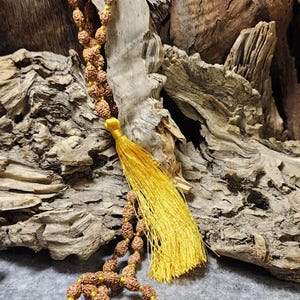 May include: A brown beaded necklace with a yellow tassel. The beads are a natural, textured material, strung together with yellow thread. The tassel is a bright, golden yellow. The necklace is displayed against a backdrop of weathered wood.
