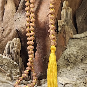 May include: A long necklace featuring numerous small, round, brown, textured rudraksha beads strung on a bright yellow cord. The necklace terminates in a single, long, vibrant yellow silk tassel that hangs from the bottom of the bead strand.