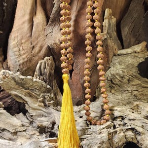 May include: A long mala necklace featuring numerous small, round, brown, textured rudraksha beads strung on a bright yellow cord. The necklace terminates in a single, long, vibrant yellow tassel that hangs from the bottom of the bead strand.