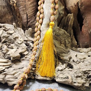 May include: A long mala necklace featuring numerous small, round, brown, textured rudraksha beads strung on a bright yellow cord. The necklace includes a prominent, long, vibrant yellow tassel hanging from the center of the bead strand.