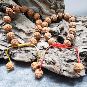 May include: Two strands of prayer beads with textured, light brown beads. One strand has a yellow cord, the other a red cord. The beads are strung on a brown leather-like material. The background is a piece of weathered wood.