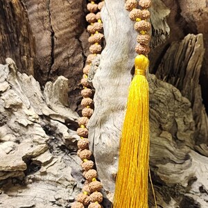May include: A long mala necklace featuring numerous small, round, brown, textured rudraksha beads strung on a bright yellow cord, finished with a long, vibrant yellow silk tassel hanging from the bottom of the bead strand.