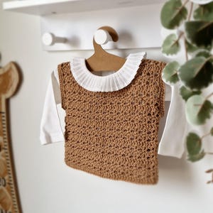May include: A brown crocheted sleeveless top with a white ruffled collar hanging on a wooden hanger.
