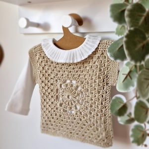 Puede incluir: Un top de ganchillo beige con cuello blanco con volantes y mangas largas, colgado de una percha de madera. El diseño de ganchillo presenta un motivo floral en el centro. El top es probablemente para un bebé o un niño pequeño.