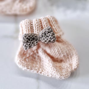 Peut inclure: Une paire de chaussons de bébé tricotés en rose clair, chacun orné d'un nœud gris. Les chaussons ont un bord côtelé et un motif de tricot texturé, parfaits pour un nouveau-né. L'arrière-plan est flou, mettant en valeur les chaussons.